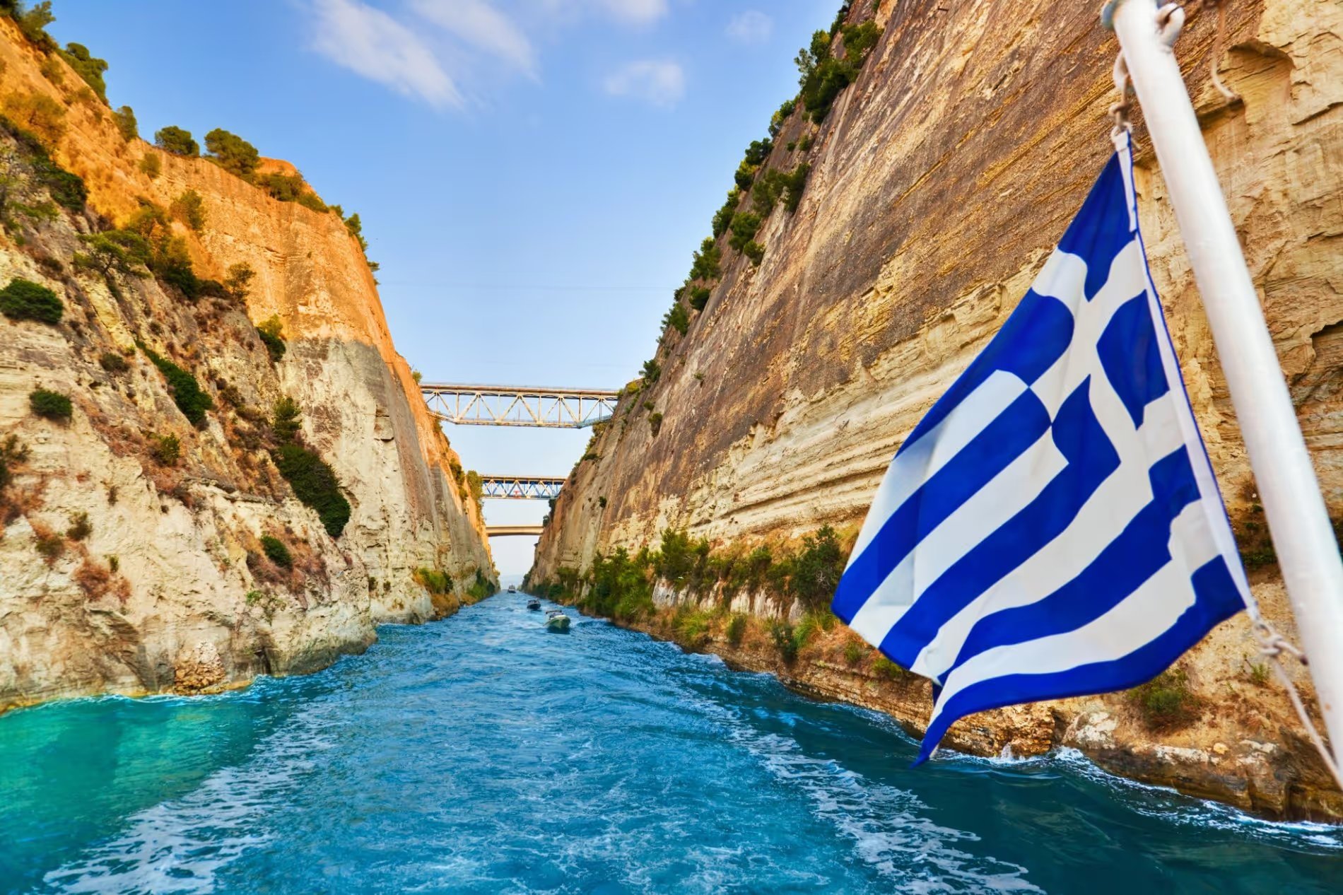 Vue du canal de Corinthe en Grèce, avec une falaise rocheuse et le drapeau grec flottant au premier plan.