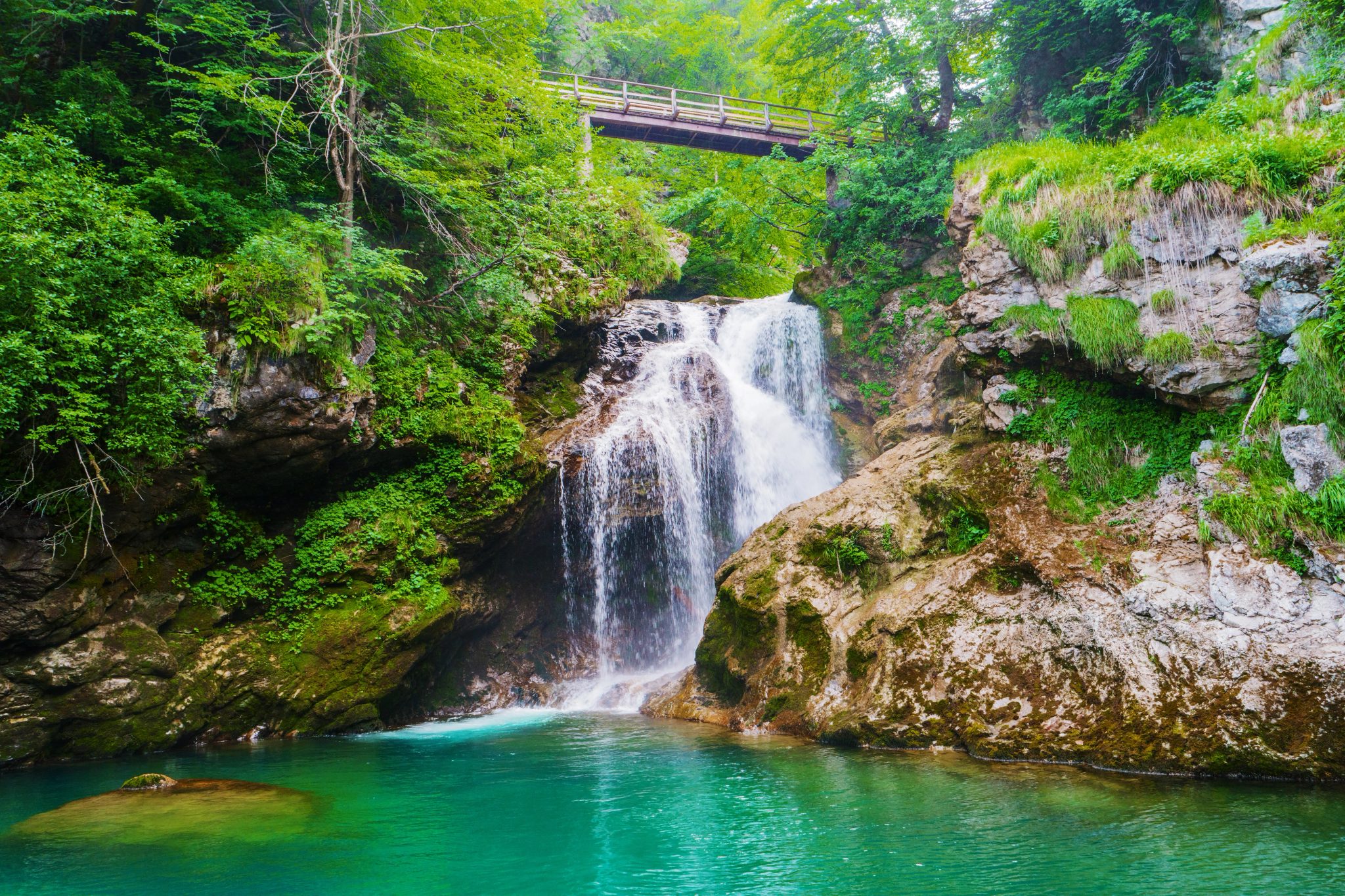 Cascade Šum et bassin au pied des rochers, avec un pont en bois en arrière-plan à Vintgar.