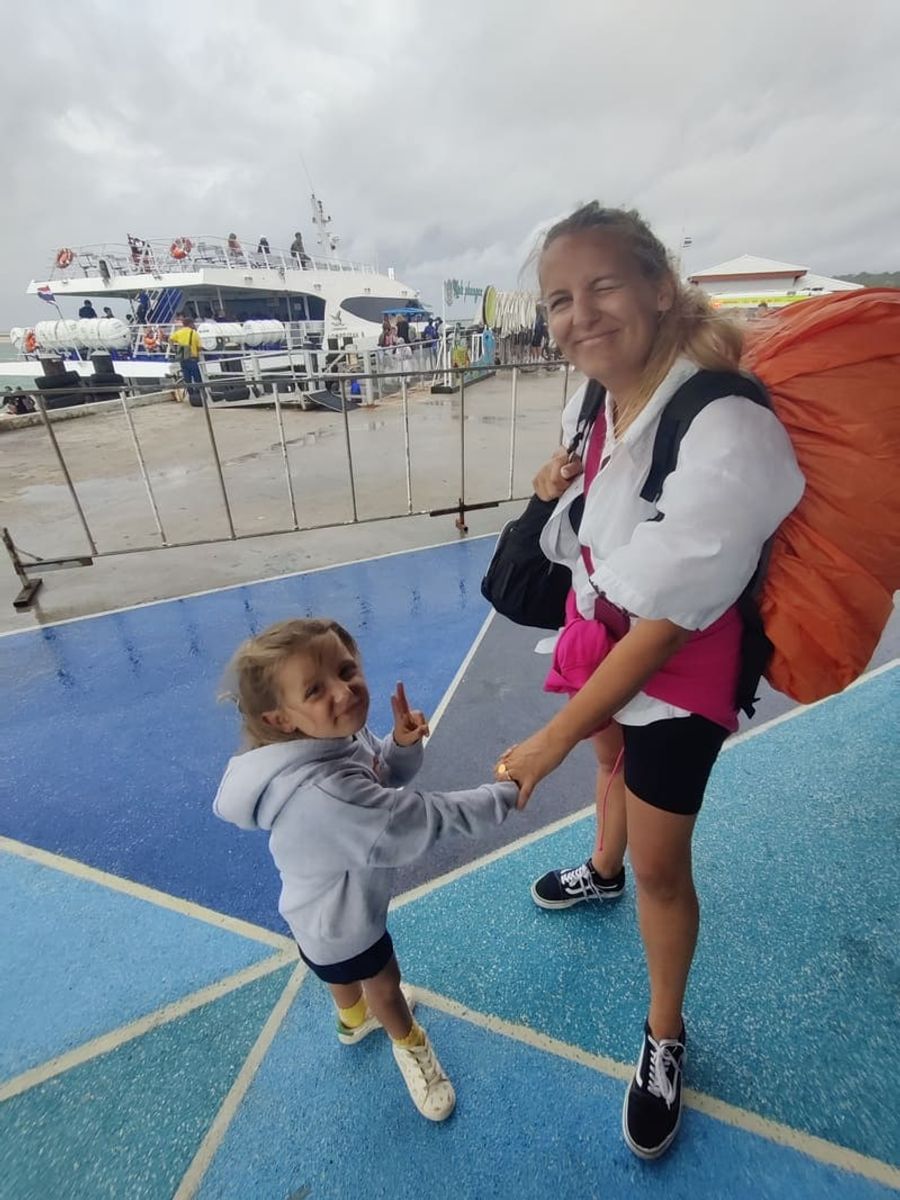 Maud et Anna souriant, tenant la main, sur un quai sous la pluie, avec un ferry en arrière-plan, prêts à embarquer à Koh Phangan.