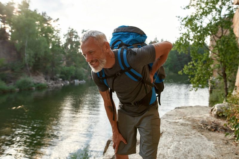 Un homme avec un sac à dos bleu souffre de maux de dos lors d'une randonnée près d'une rivière entourée de végétation.