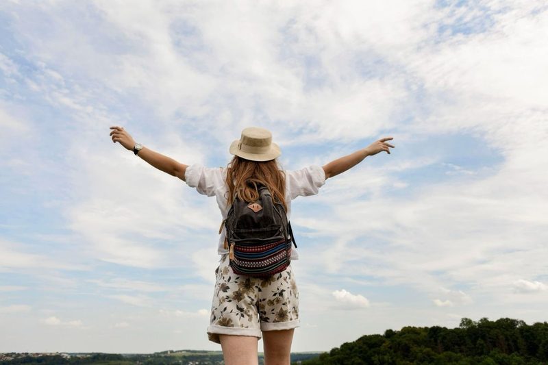 Voyageuse avec un chapeau et un sac à dos admirant la nature les bras ouverts, symbole de liberté et de voyage écoresponsable.