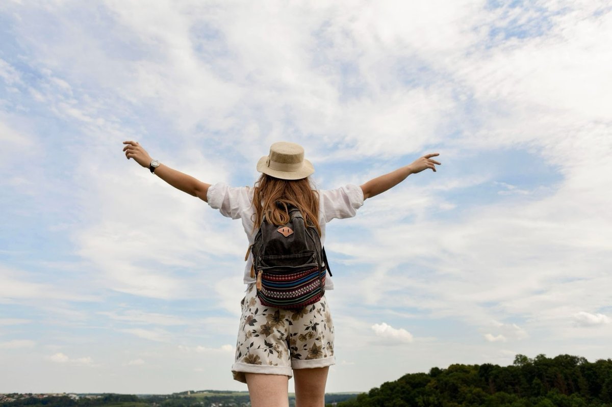 Voyageuse avec un chapeau et un sac à dos admirant la nature les bras ouverts, symbole de liberté et de voyage écoresponsable.