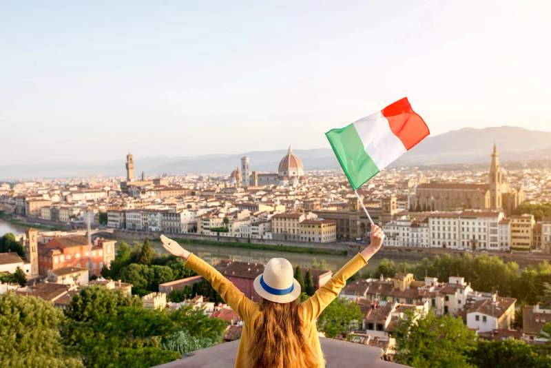 Une personne de dos, bras levés, brandissant un drapeau italien face à une vue panoramique de Florence, en Italie