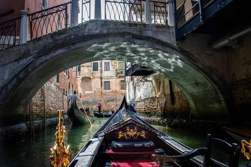 Une gondole navigue sous un pont dans un canal pittoresque de Venise, Italie, avec des reflets de lumière sur l'eau et les murs en briques environnants.