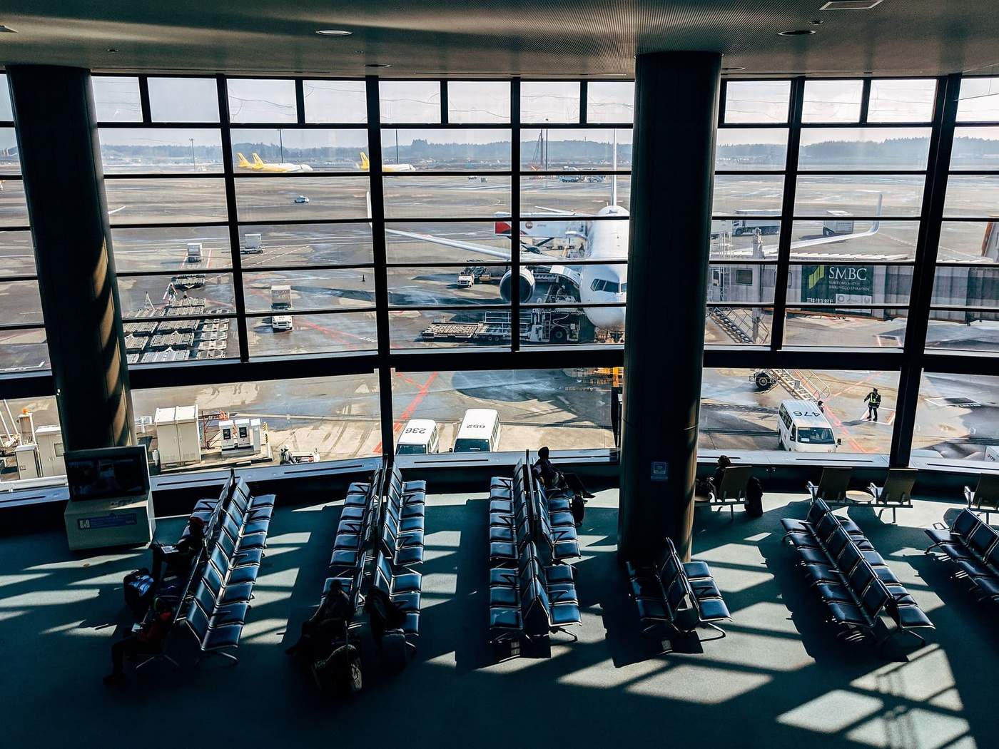 Vue intérieure d’un terminal d’aéroport avec des rangées de sièges vides et un avion stationné à une passerelle d’embarquement. Quelques voyageurs sont assis, observant les avions à l’extérieur à travers les grandes baies vitrées.