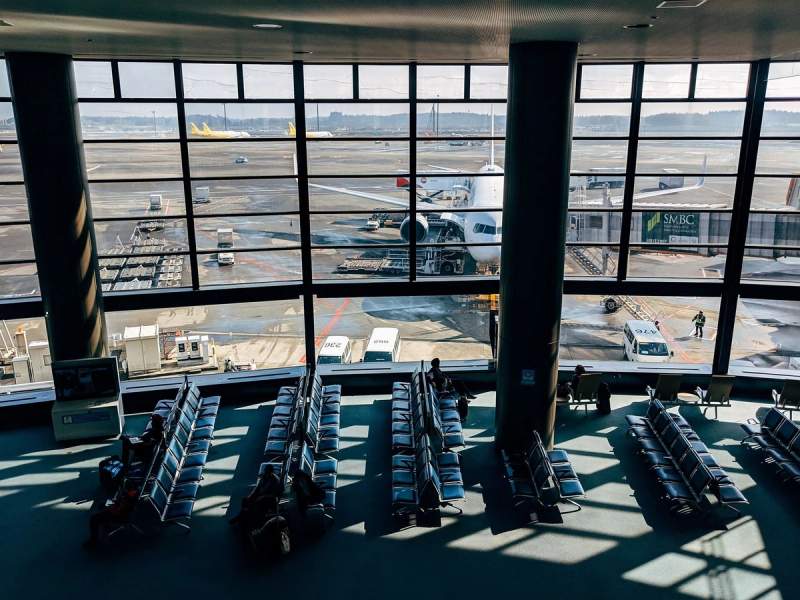 Vue intérieure d’un terminal d’aéroport avec des rangées de sièges vides et un avion stationné à une passerelle d’embarquement. Quelques voyageurs sont assis, observant les avions à l’extérieur à travers les grandes baies vitrées.