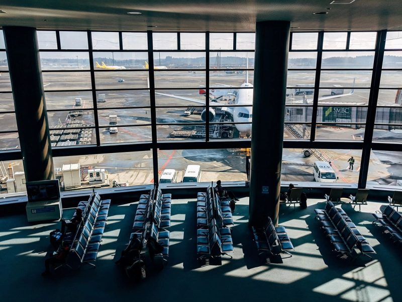 Vue intérieure d’un terminal d’aéroport avec des rangées de sièges vides et un avion stationné à une passerelle d’embarquement. Quelques voyageurs sont assis, observant les avions à l’extérieur à travers les grandes baies vitrées.