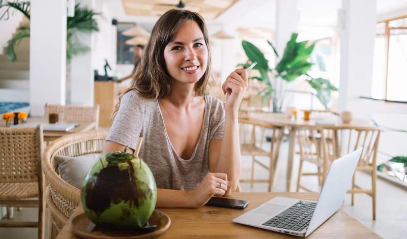 Jeune femme souriante travaillant sur un ordinateur portable dans un café, avec une noix de coco fraîche à côté d’elle sur la table.