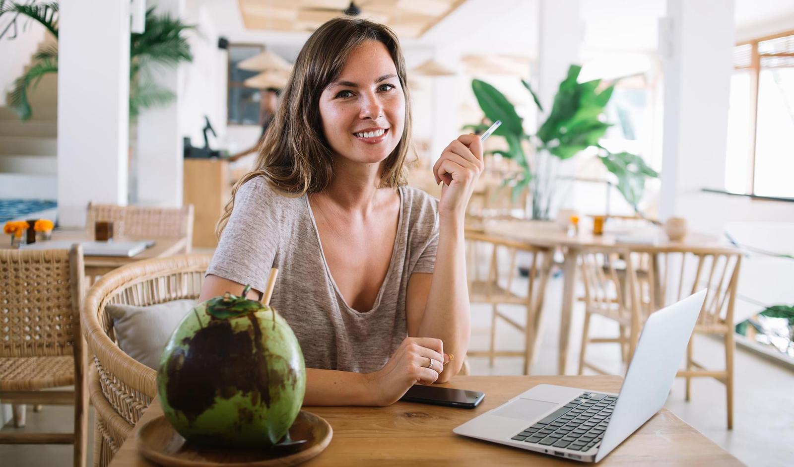 Jeune femme souriante travaillant sur un ordinateur portable dans un café, avec une noix de coco fraîche à côté d’elle sur la table.