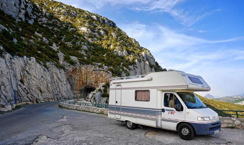 Un camping-car garé sur une route de montagne près d'un tunnel, sous un ciel bleu clair.