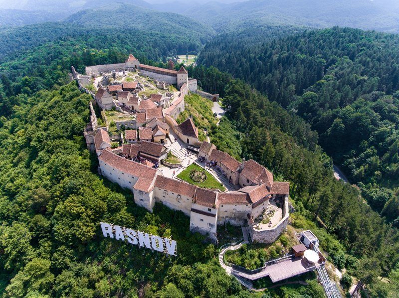 Vue aérienne de la forteresse médiévale entourée de forêts, avec des remparts et des toits en tuiles rouges.