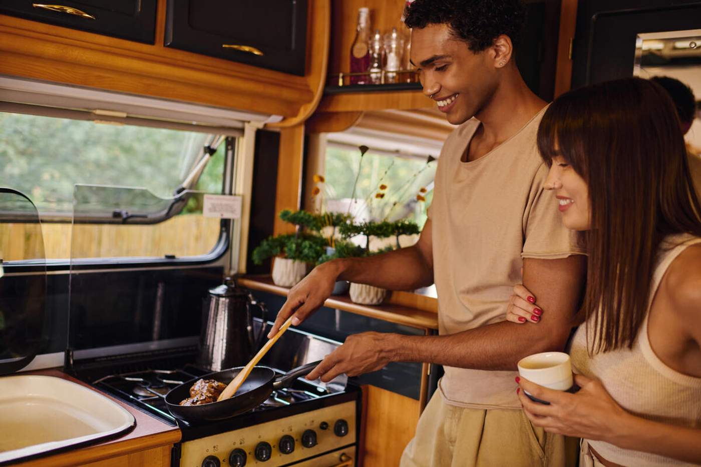 Couple préparant un repas dans la cuisine d'un camping-car, l'homme cuisine à la poêle sur une plaque gaz pendant que sa compagne tient une tasse de café