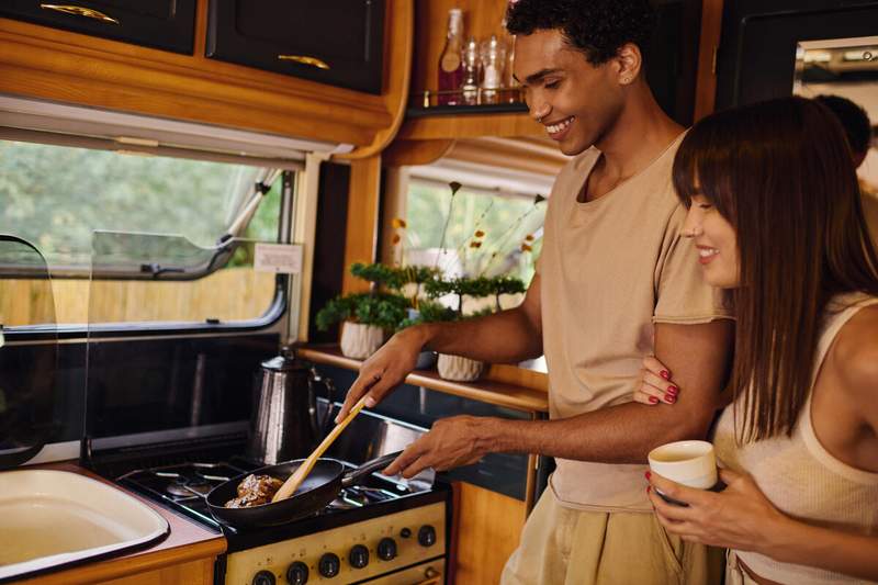 Couple préparant un repas dans la cuisine d'un camping-car, l'homme cuisine à la poêle sur une plaque gaz pendant que sa compagne tient une tasse de café
