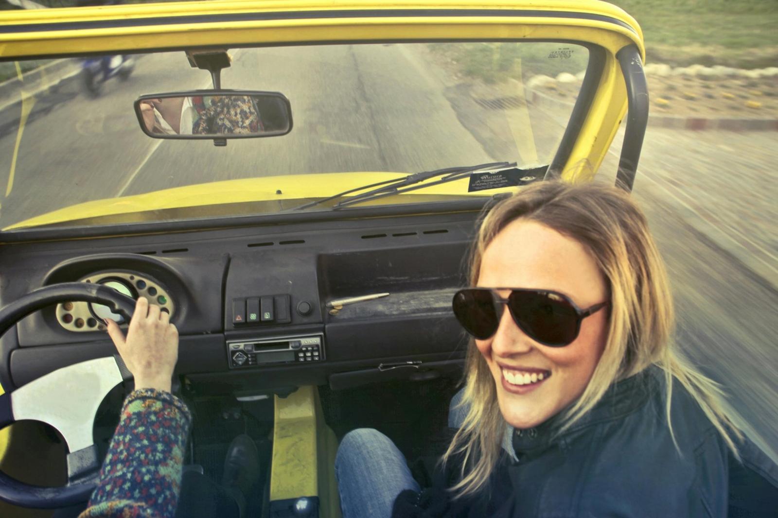 Scène de road trip avec une femme souriante portant des lunettes de soleil assise à l’avant d’une voiture jaune, tandis qu’une autre personne conduit.