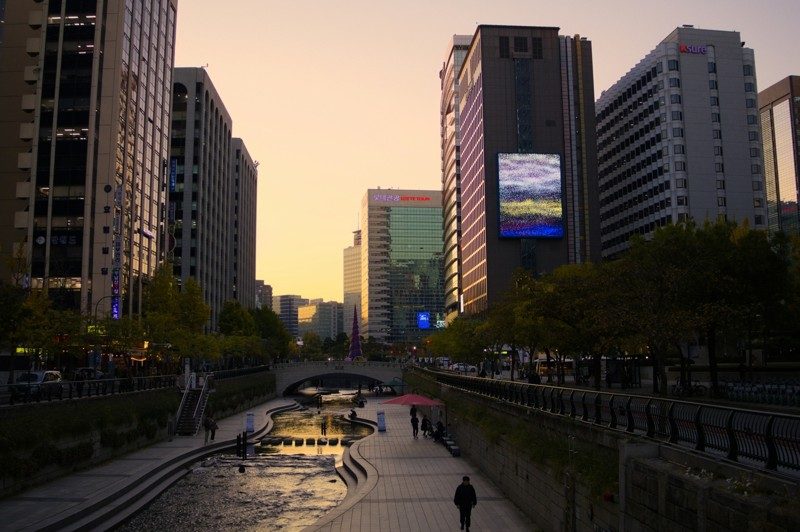 Promenade le long du ruisseau Cheonggyecheon à Séoul au coucher du soleil, bordée de gratte-ciel et d’écrans lumineux.