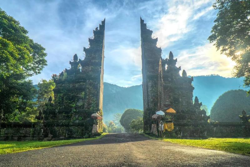 Portail Handara à Bali : deux grandes colonnes sculptées encadrent une route menant à des montagnes enveloppées de brume, avec des offrandes traditionnelles balinaises au pied.