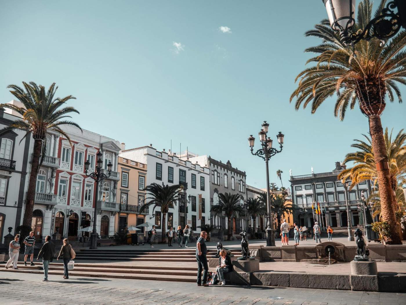 Place ensoleillée en Espagne avec des bâtiments historiques et des palmiers, des personnes se promenant et profitant du cadre paisible.
