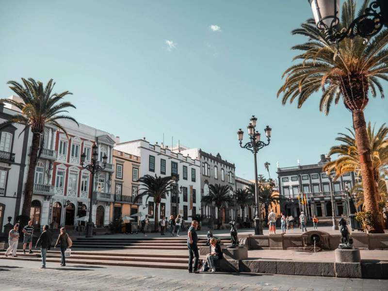 Place ensoleillée en Espagne avec des bâtiments historiques et des palmiers, des personnes se promenant et profitant du cadre paisible.