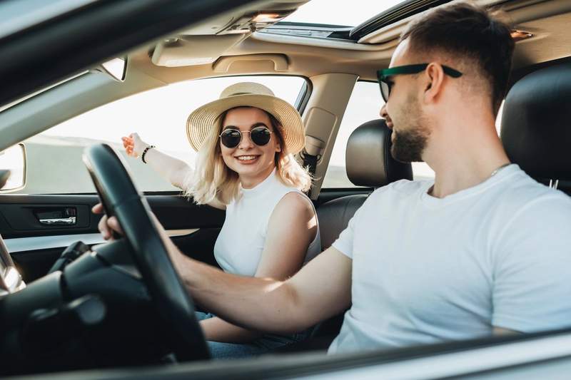 Couple souriant en voiture pendant un voyage, la femme portant un chapeau et des lunettes de soleil, l’homme au volant.