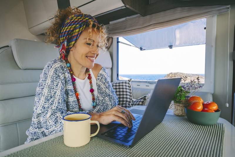 Femme nomade numérique travaillant sur un ordinateur portable dans un van face à la mer, tasse de café et fruits frais à ses côtés.