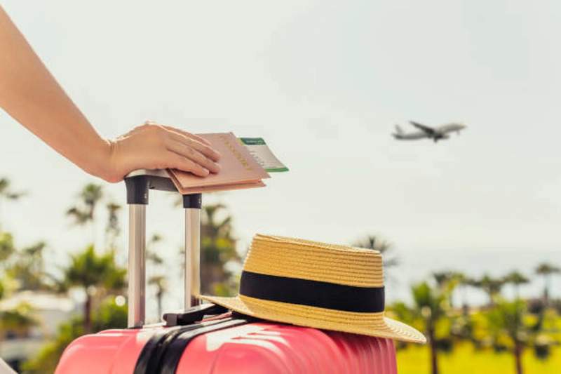 ue rapprochée d'une valise rose avec un chapeau de paille posé dessus, une main tenant un passeport et une carte d'embarquement, avec un avion décollant en arrière-plan et des palmiers flous sous un ciel ensoleillé.