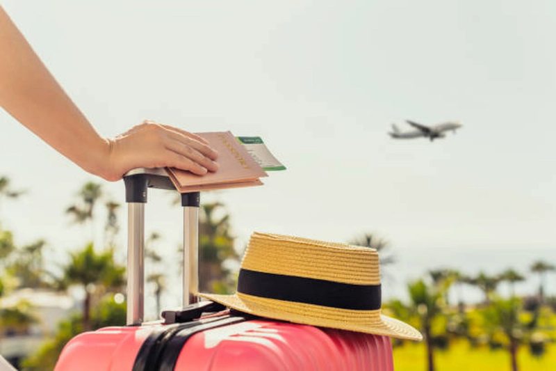 ue rapprochée d'une valise rose avec un chapeau de paille posé dessus, une main tenant un passeport et une carte d'embarquement, avec un avion décollant en arrière-plan et des palmiers flous sous un ciel ensoleillé.
