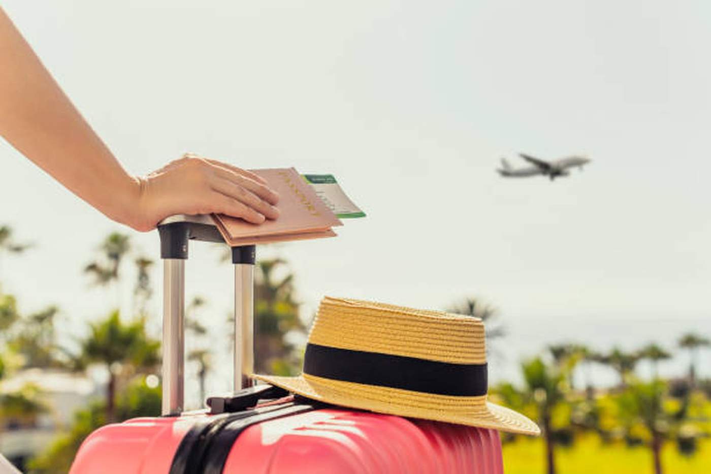 ue rapprochée d'une valise rose avec un chapeau de paille posé dessus, une main tenant un passeport et une carte d'embarquement, avec un avion décollant en arrière-plan et des palmiers flous sous un ciel ensoleillé.