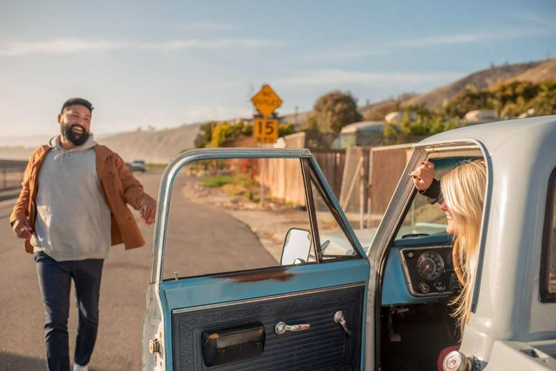 Couple souriant à côté d'un pick-up vintage bleu et blanc sur une route de bord de mer