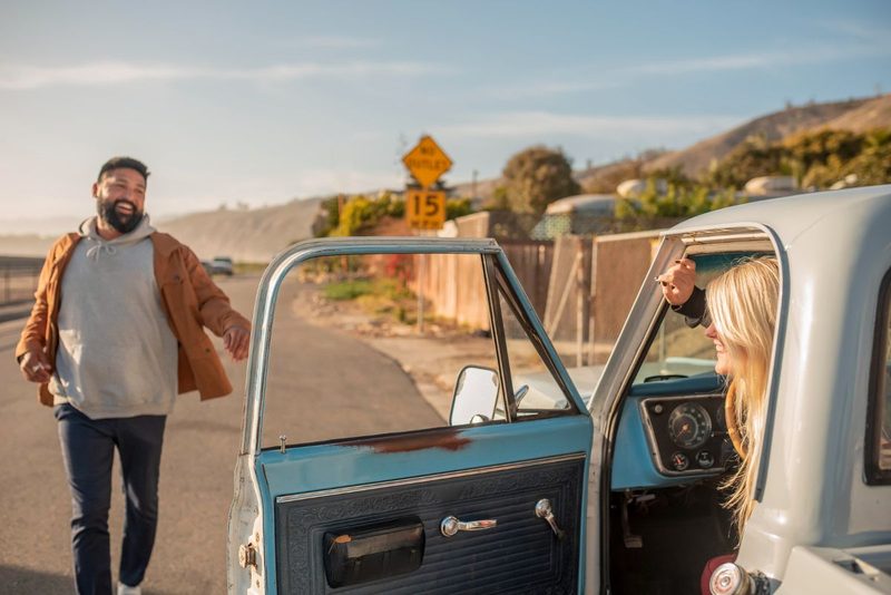 Couple souriant à côté d'un pick-up vintage bleu et blanc sur une route de bord de mer