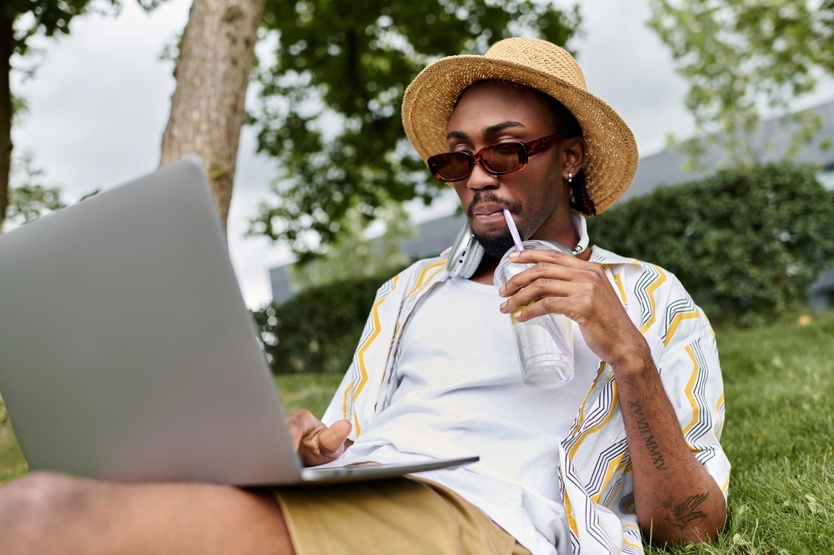 Digital nomade travaillant sur son laptop assis dans l'herbe en extérieur, avec un chapeau de paille et des écouteurs autour du cou