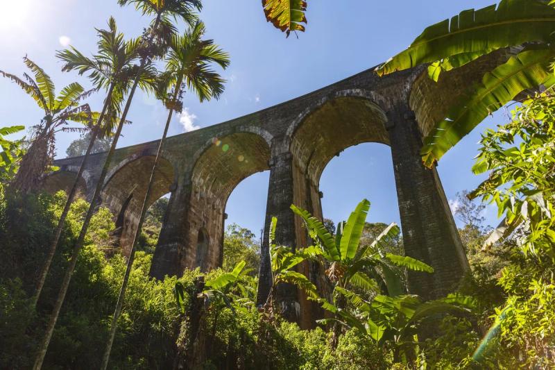 Vue en contre-plongée du Nine Arch Bridge à Ella, Sri Lanka, entouré de végétation tropicale et de bananiers sous un ciel bleu.