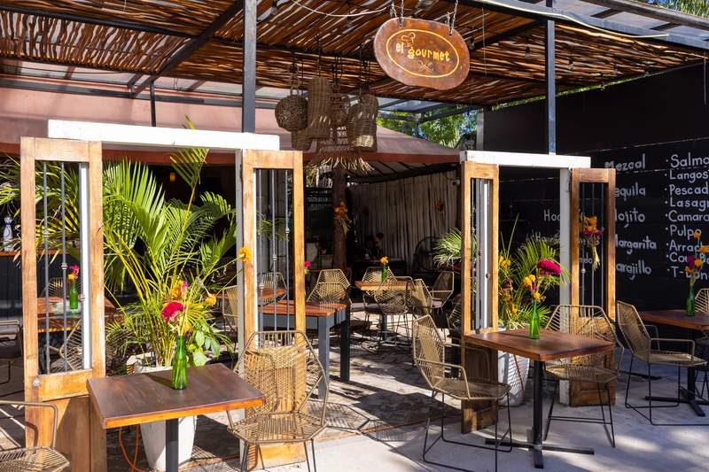 Terrasse ensoleillée du restaurant El Gourmet à Tulum, avec chaises en rotin, tables en bois et plantes tropicales décoratives.