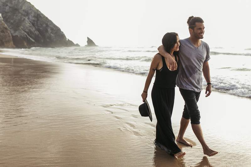 Couple se promenant pieds nus sur une plage sauvage au coucher du soleil, au bord de falaises rocheuses, lors d'un road trip en Europe
