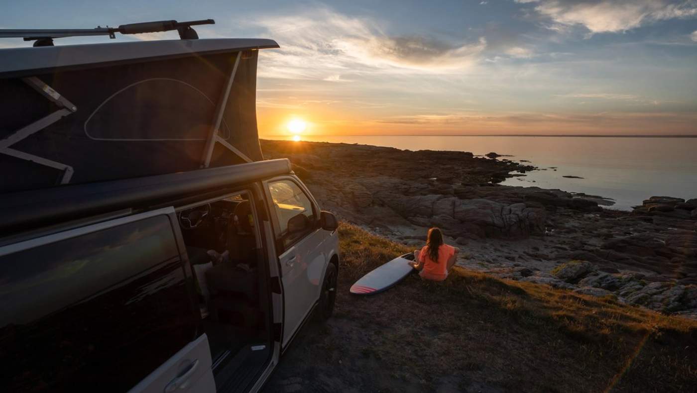 Van aménagé stationné en bord de mer au coucher du soleil, avec une voyageuse assise face à l’océan et une planche de surf, ambiance road trip et vanlife.
