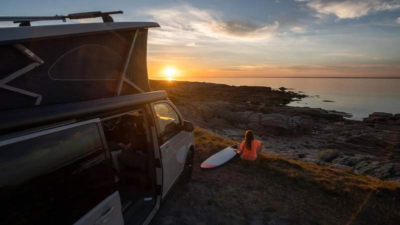 Van aménagé stationné en bord de mer au coucher du soleil, avec une voyageuse assise face à l’océan et une planche de surf, ambiance road trip et vanlife.