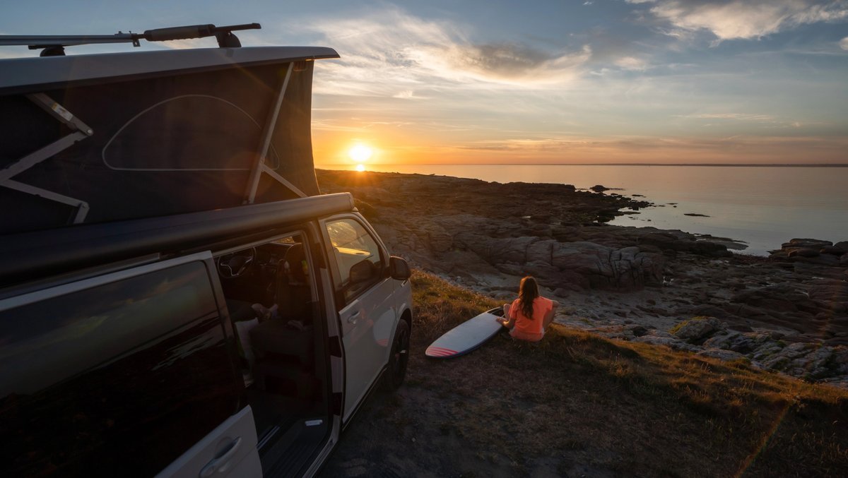 Van aménagé stationné en bord de mer au coucher du soleil, avec une voyageuse assise face à l’océan et une planche de surf, ambiance road trip et vanlife.