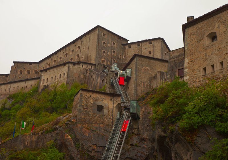 Vue du Fort Bard, une imposante forteresse perchée sur une colline, entourée de verdure et accessible par un funiculaire.