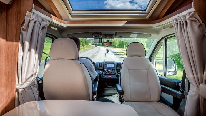 Vue depuis l’intérieur d’un van aménagé roulant sur une route de campagne, avec toit panoramique et sièges avant beiges.