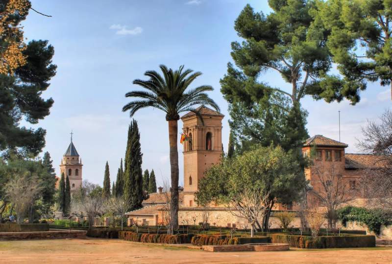 Vue sur les tours et les jardins de l'Alhambra à Grenade, entourée de palmiers et de cyprès sous un ciel dégagé.