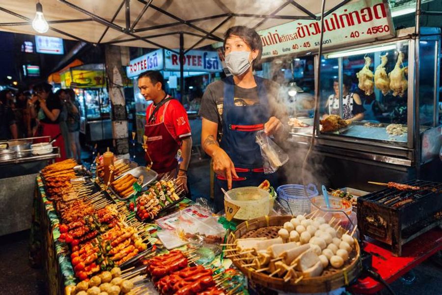 Scène animée du marché de nuit de Chiang Mai, Thaïlande, avec un vendeur servant des brochettes colorées et des stands illuminés.