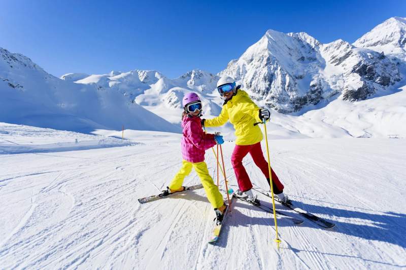 Deux personnes en tenue de ski colorée sur une piste enneigée avec de majestueuses montagnes en arrière-plan.