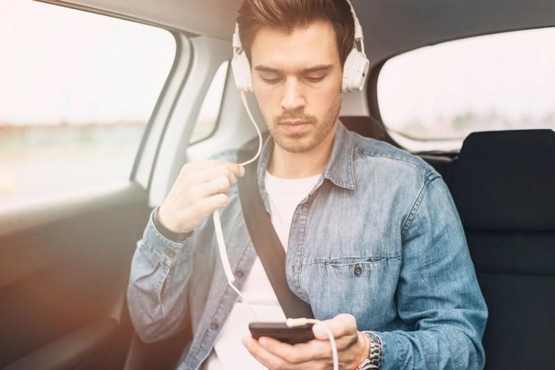 Un homme assis à l’arrière d’une voiture écoute de la musique avec un casque blanc, regardant son téléphone.