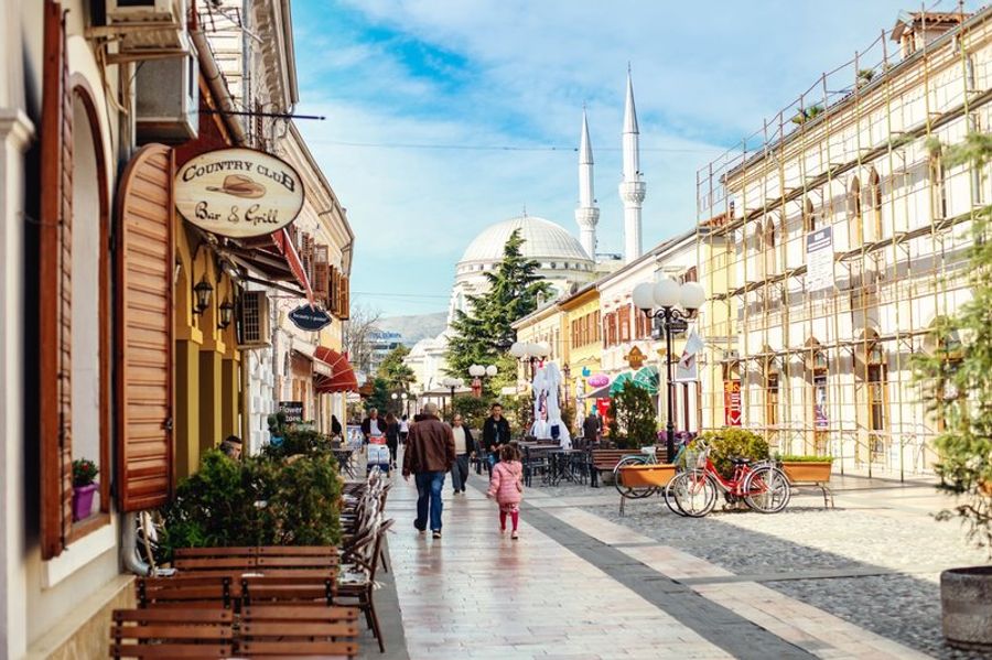 Rue pavée animée de Shkodër, bordée de cafés, avec une mosquée en arrière-plan sous un ciel bleu.