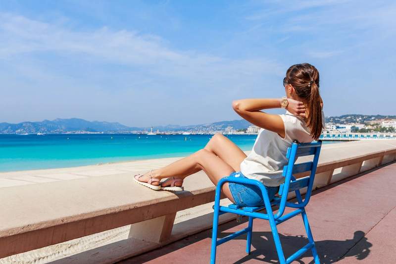 Femme de dos assise sur une chaise bleue, admirant la mer turquoise depuis la promenade de la Côte d’Azur par une journée ensoleillée.