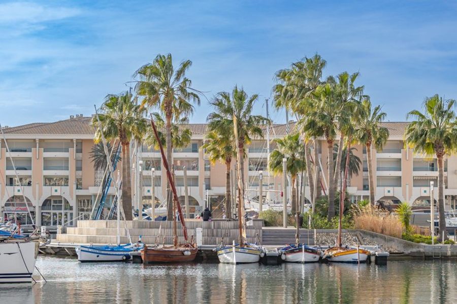 Vue sur le port de Fréjus en France avec des bateaux amarrés devant des palmiers et un immeuble moderne sous un ciel bleu clair.