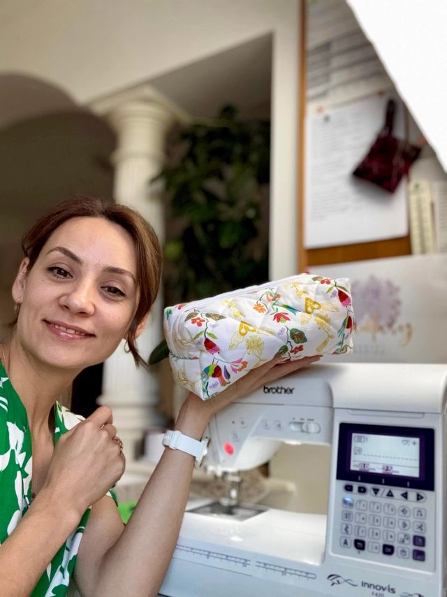 Kristina smiling, holding up a boxy quilted pouch in a bright floral print with a white zip, beside her sewing machine