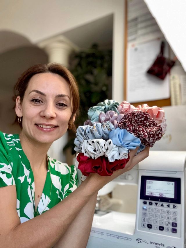 Kristina smiling beside her sewing machine, holding up an armful of scrunchies in satin, florals and polka dots