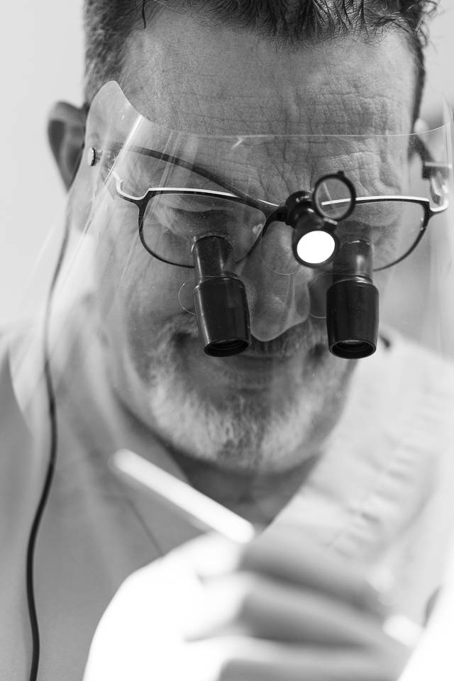 black and white close up photo of dentist wearing protective face mask and loupes examining patient in surgery