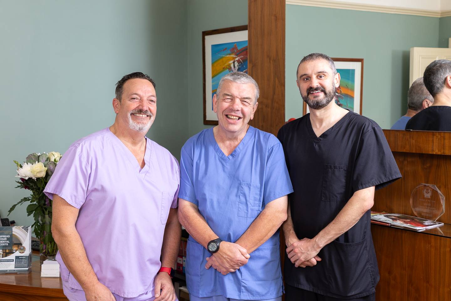 Three dentist in purple, blue and black scrubs laughing together at the camera