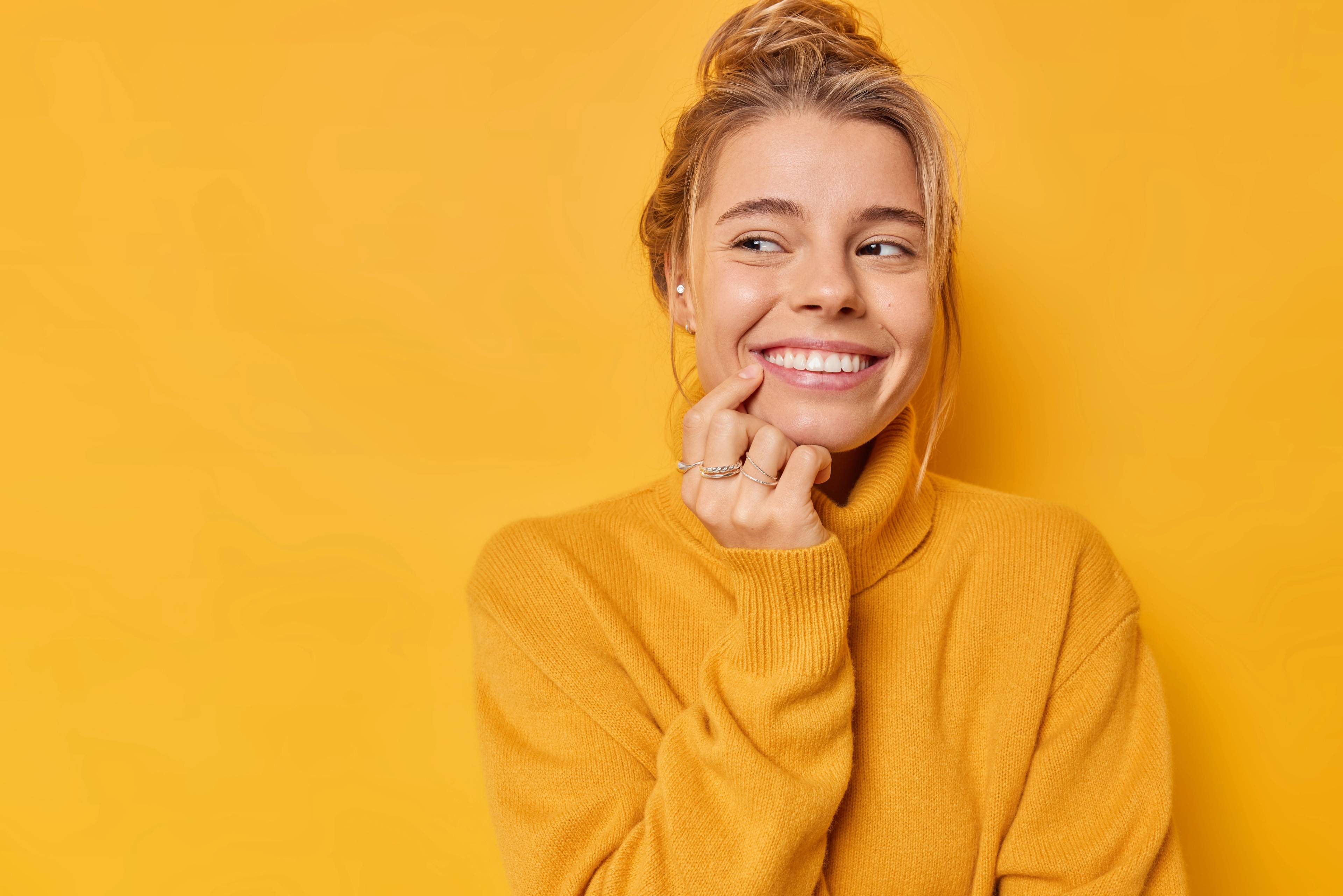 female wearing yellow jumper standing in front of yellow background touching teeth and smiling looking to the side
